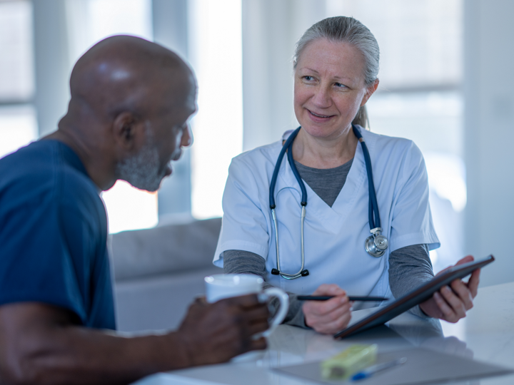A female homecare nurse sits with a male patient at the kitchen table as they meet to discuss his health concerns. The nurse is wearing white scrubs and holding out tablet as the two talk.