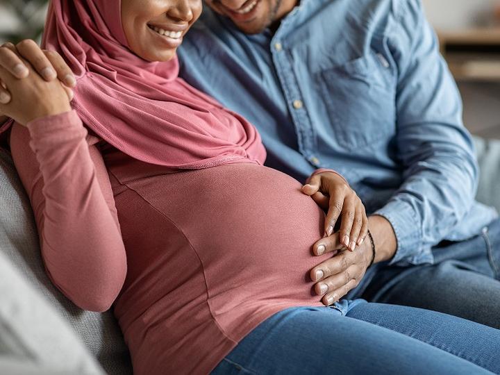 Loving husband tenderly touching belly of his pregnant Muslim wife while sitting together on couch in living room.