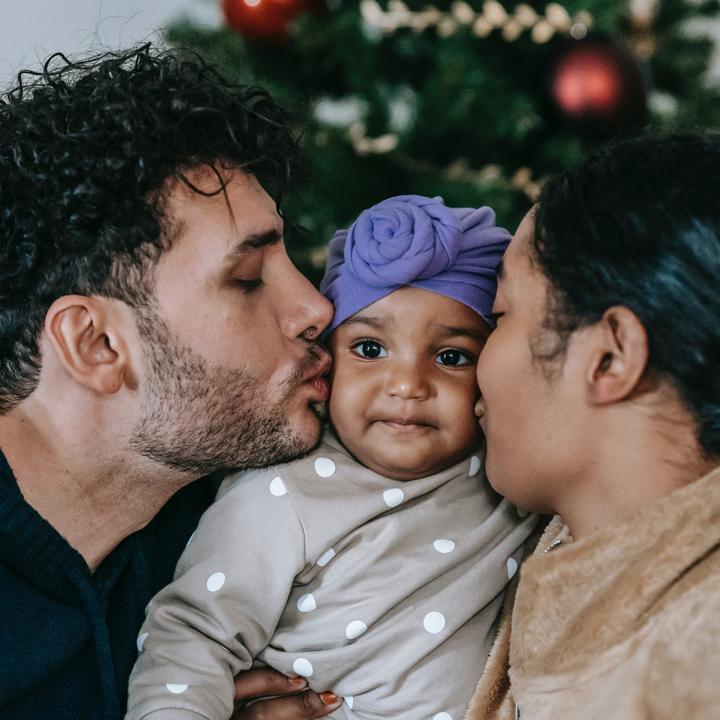 Parents kissing baby girl's cheeks with festive wreath in the background.